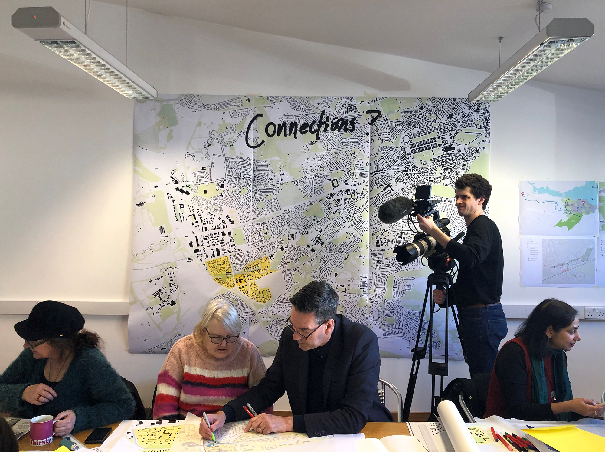 Two people sit at a table sketching on a large piece of paper. There is a person with a video camera in the background, and a large map pinned to the wall.