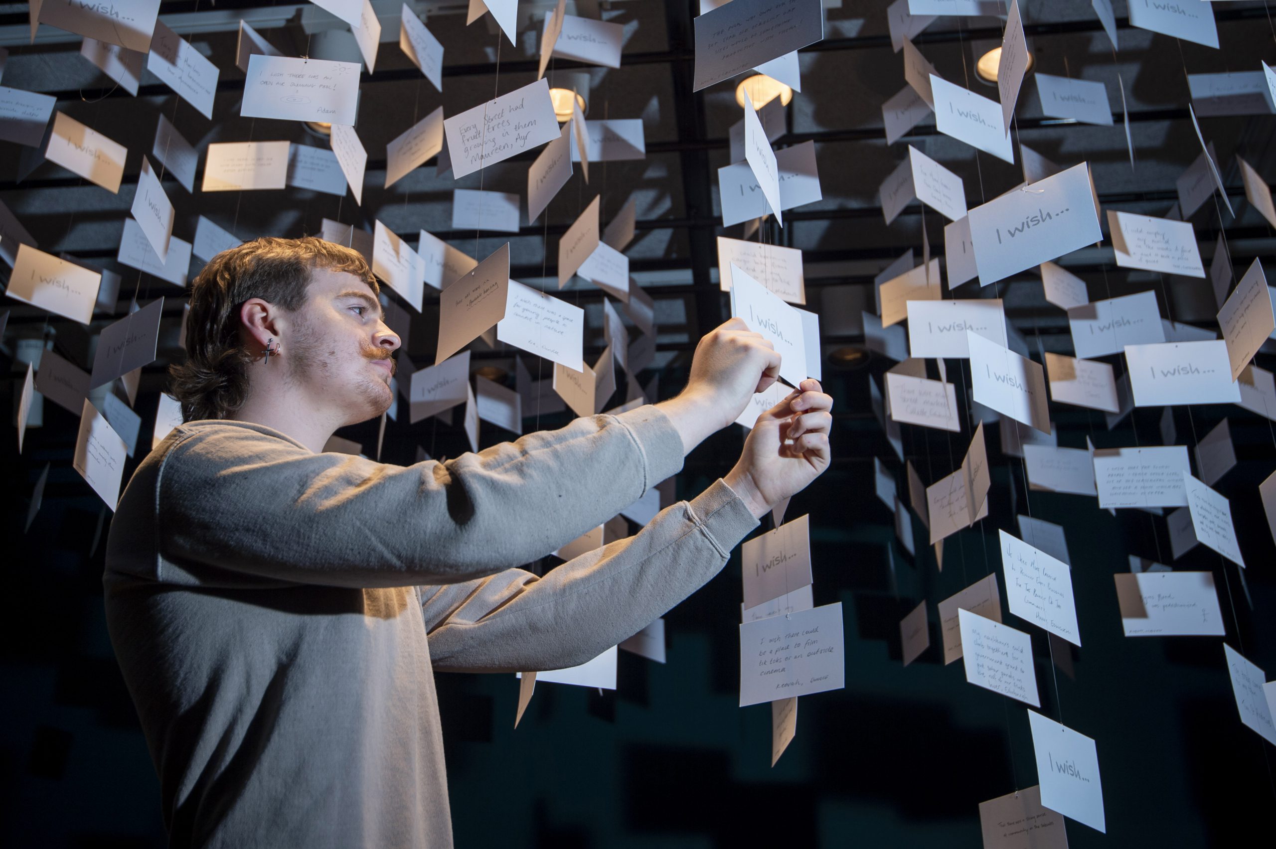 A man holds a post card standing in a "cloud" of white cards suspended from strings in a dark room.