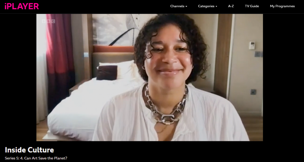 Image is of artist Alberta Whittle smiling to camera. She is wearing a white shirt with a chunky chain necklace in silver.