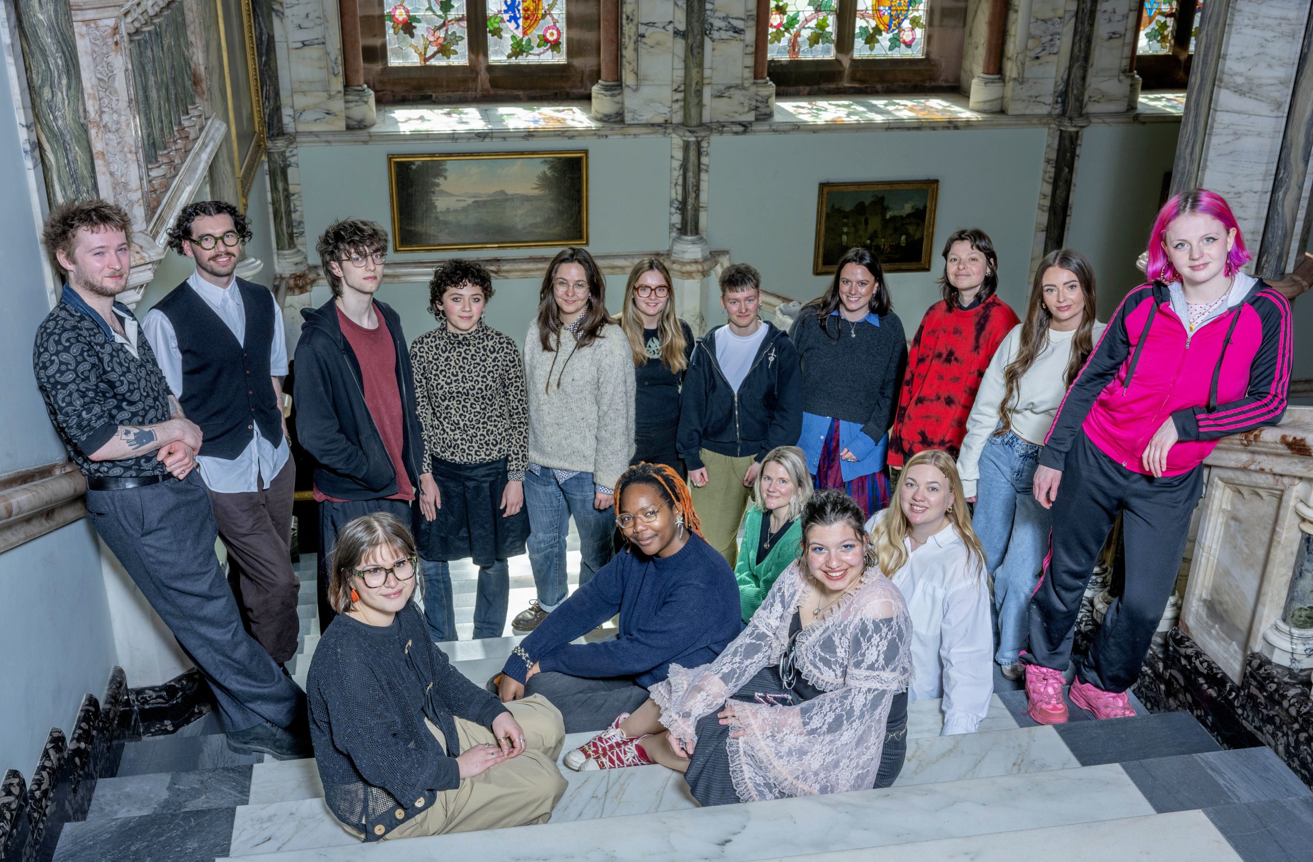 A group of people posed looking at the camera on a set of marble stairs with stained glass windows in the backdrop.